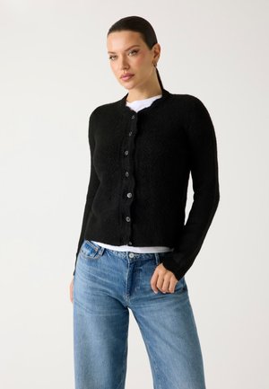 Kobieta z związanymi włosami, ubrana w czarny sweter zapinany na guziki, pod który ma białą koszulę, oraz jasnoniebieskie dżinsy, na jednolitym tle.