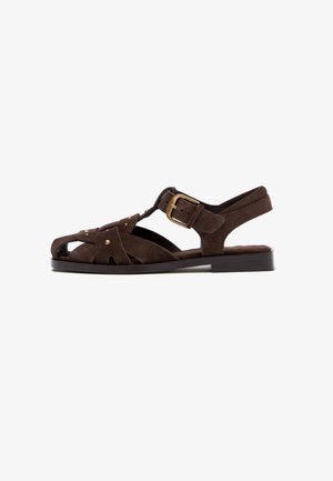 Sandalo piatto in suede marrone scuro con punta aperta, design a T, fibbia dorata e piccoli borchietti decorativi sulle cinghie laterali.