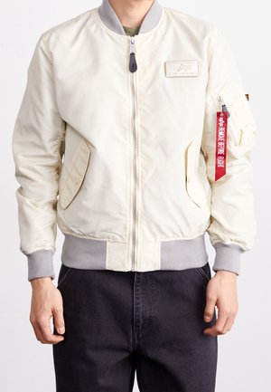 Personne portant une veste bombers crème avec des poignets et une ceinture gris, une fermeture éclair à l’avant, des poches à rabat et une étiquette rouge "Remove Before Flight" sur la fermeture éclair de la manche.