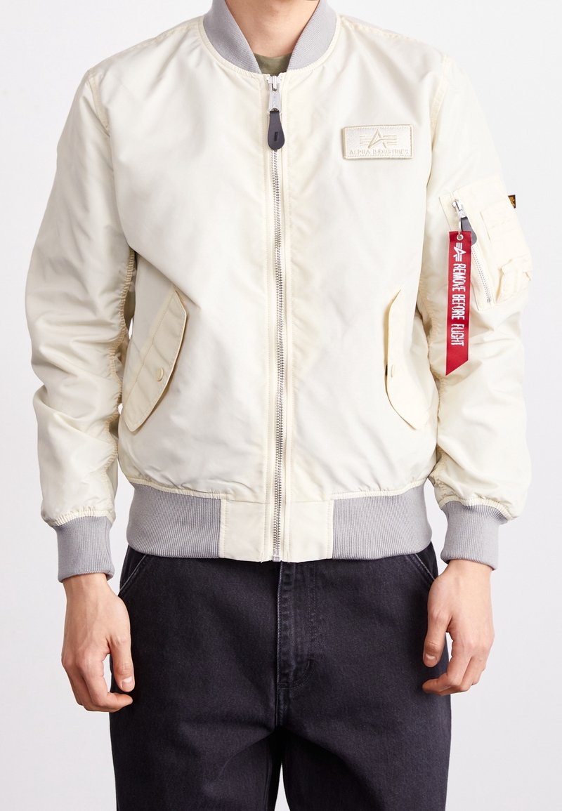 Personne portant une veste bombers crème avec des poignets et une ceinture gris, une fermeture éclair à l’avant, des poches à rabat et une étiquette rouge "Remove Before Flight" sur la fermeture éclair de la manche.
