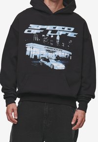 Svart hoodie med grafiskt tryck av en bil och en bensinstation. Kängurufickor, ribbad ärmslut och nederkant, avslappnad passform.