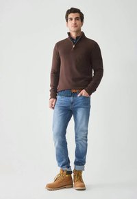 Pulôver castanho de malha com fecho de correr, jeans azul claro e botas de trabalho castanhas. Parte superior texturada e punhos canelados. Roupa casual para atividades ao ar livre.