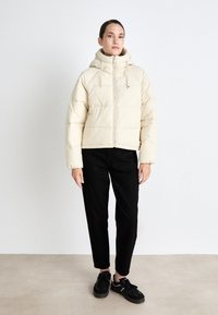 Beige puffer jacket with a hood, featuring a front zipper, quilted design, and elastic cuffs, paired with black pants and sneakers.