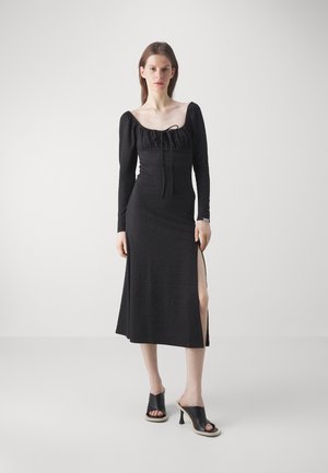 Black long-sleeve dress with ruched bodice, scoop neckline, front tie detail, and side slit; paired with black open-toe block-heeled shoes.