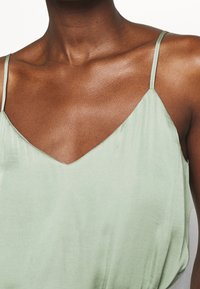 Grön sidenblus med V-ringning och tunna axelband, som har en slät textur och subtil lyster. Minimalistisk design utan mönster.