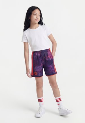 Jeune fille debout avec une main dans la poche, portant un t-shirt blanc, un short de sport violet-rouge, des chaussettes blanches à rayures rouges, et des baskets blanches.