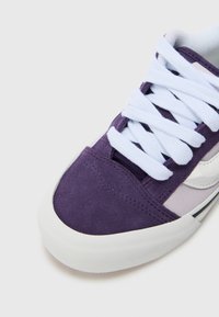 Adidași din suede mov cu șireturi albe, având un vârf texturat și detalii cu logo pe lateral, pe un fundal gri deschis.