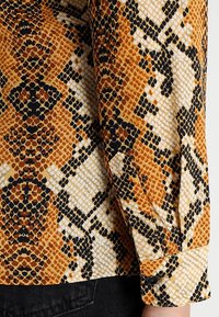 Blusa a maniche lunghe con motivo a stampa serpente nei toni di marrone, nero e crema. Realizzata in tessuto morbido con dettagli sui polsini abbottonati.