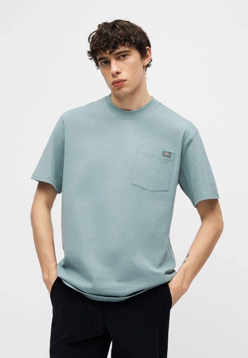 Giovane con capelli ricci che indossa una maglietta azzurra a maniche corte con taschino sul petto e pantaloni neri, con le mani nelle tasche.