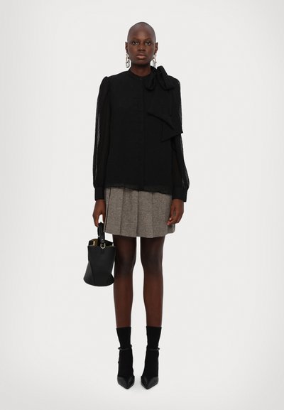 Schwarze, strukturierte Bluse mit einer Schleifenapplikation, kombiniert mit einem plissierten karierten Rock. Die Accessoires umfassen eine schwarze Bucket-Bag und spitze Schuhe.