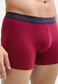 Boxer bóxer burdeos para hombre con una cinturilla azul marino que presenta el logotipo "TOM TAILOR" en rojo. Textura suave y diseño ajustado.