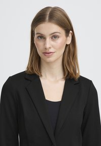 Nuori nainen, jolla on suorat vaaleanruskeat hiukset, pukeutuneena mustaan bleiseriin ja tummaan yläosaan, katsoo eteenpäin yksinkertaista vaaleaa taustaa vasten.