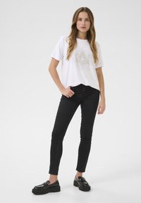 T-shirt blanc à manches courtes avec un design graphique gris détaillé, associé à un pantalon noir ajusté et des mocassins noirs à semelles épaisses.