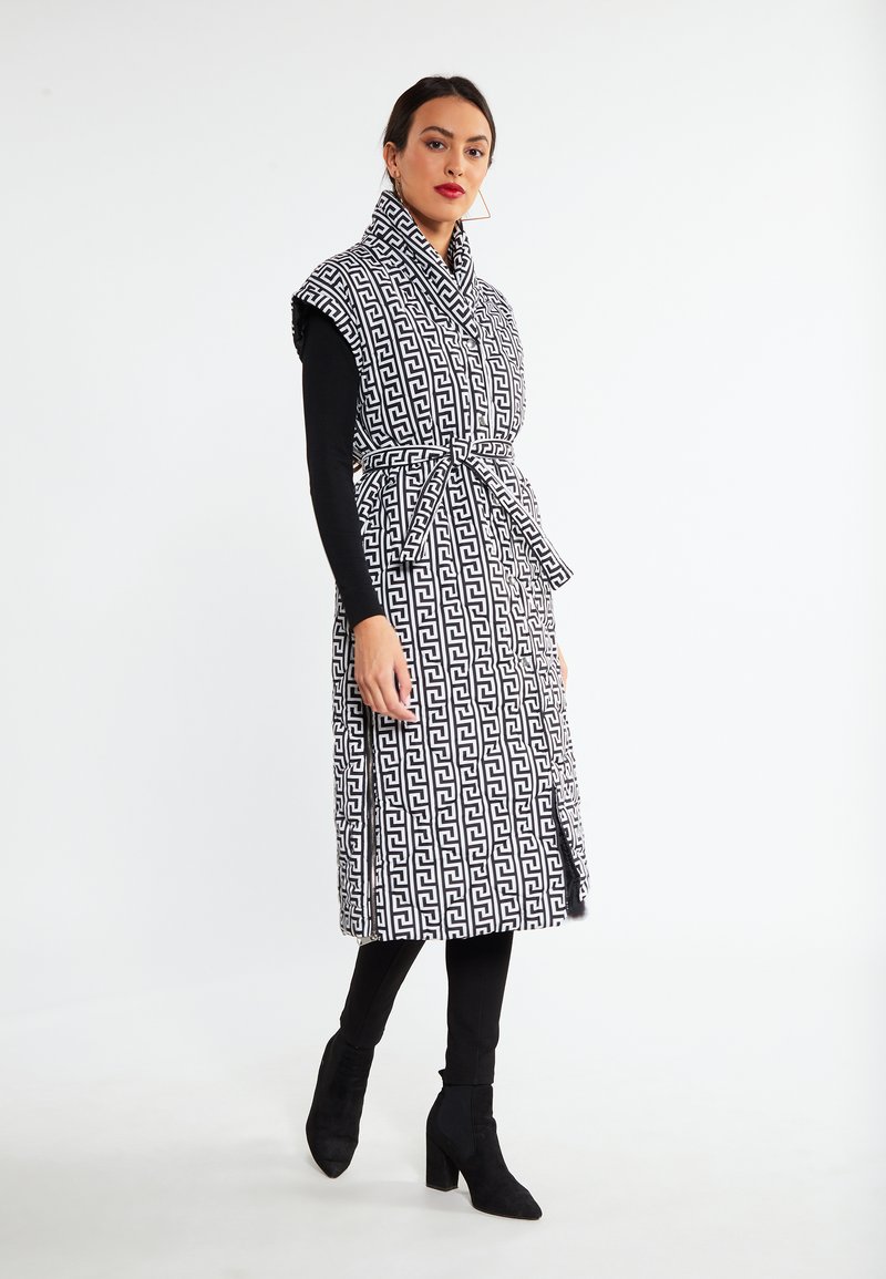 Zwart-wit gestreepte mouwloze jas met een hoge kraag, een riem in de taille en zijsplitten, gecombineerd met strakke zwarte broek en enkelboots.