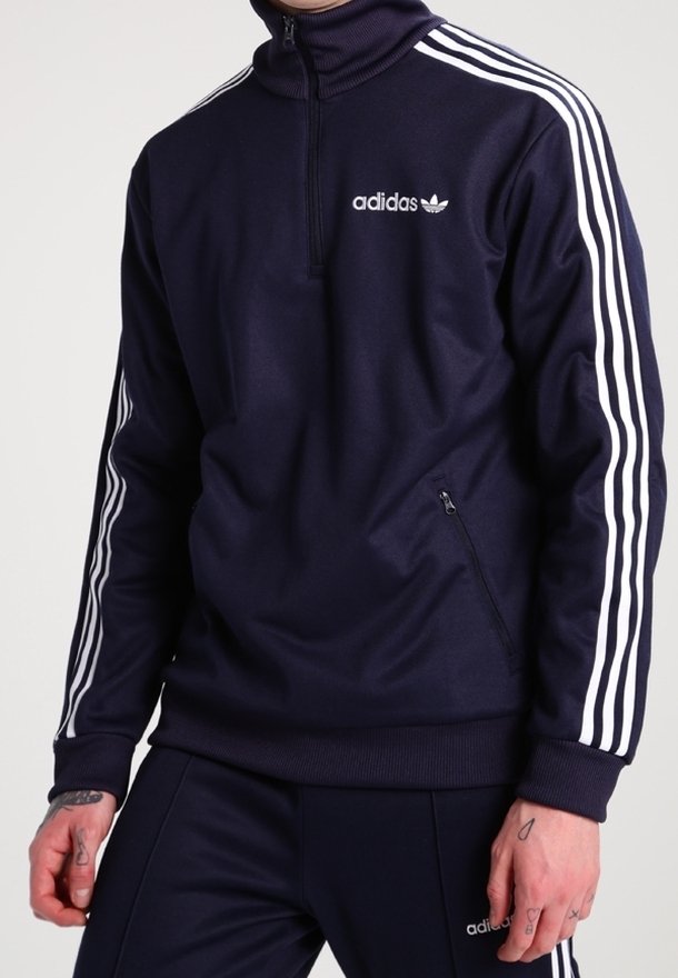 Sudadera de Adidas azul marino con alto cuello, cremallera de cuarto, acentos de tres rayas blancas en las mangas y bolsillo con cremallera lateral. Textura de tela suave.