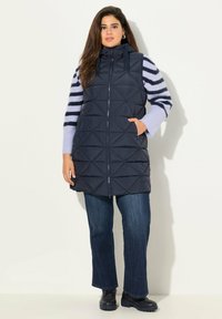 Gilet imbottito blu navy con cappuccio, caratterizzato da un motivo a rombi e chiusura frontale con zip. Abbinato a un maglione a righe e jeans scuri.