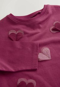 Hosszú ujjú, magenta színű pamut póló, áttetsző, rózsaszín szív applikációkkal pöttyös mintával. Lekerekített nyakvonal és látható megerősített varrások.