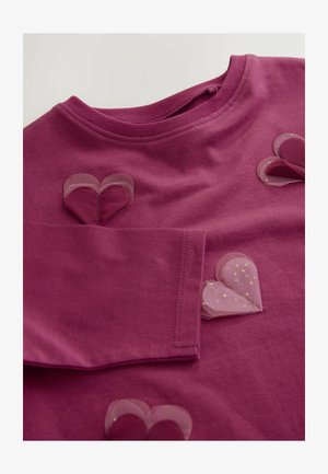 Camisa de manga larga de algodón en magenta, con apliques de corazón transparentes en rosa con lunares. Escote redondeado y costuras reforzadas visibles.