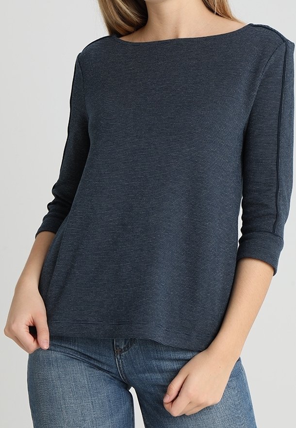 Femme portant un haut texturé bleu foncé à manches longues avec un col bateau et un jean bleu, debout les mains près des poches.