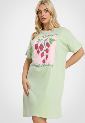 Blonde Frau trägt ein hellgrünes Kleid mit kurzen Ärmeln, das ein pinkfarbenes Stoffstück mit roten Erdbeeren und dem Schriftzug "Fruit Market Fresh Strawberries" zeigt.