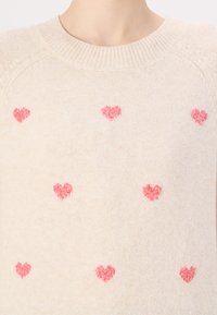 Pull en tricot beige avec un col rond, présentant des motifs de cœurs roses éparpillés. Texture douce, avec une coupe légèrement ample et des poignets côtelés.