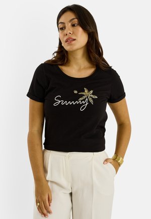 Femme portant un t-shirt noir « Sunny » avec un motif de palmier, un pantalon blanc, un bracelet en or et des bagues, debout avec une main dans la poche.