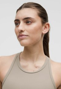 Beige urheilullinen tanktoppi, jossa on korkea kaula-auko ja leveät hihnat, ominaisuuksia sileä, joustava materiaali ja minimalistinen muotoilu.