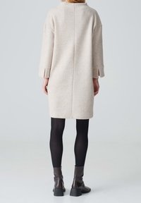 Manteau en laine beige clair avec manches longues, fentes latérales et silhouette droite, assorti à des collants noirs et des bottines marron.