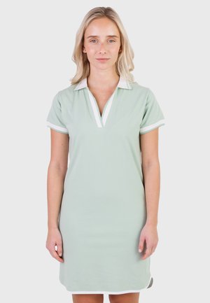 Donna bionda in piedi con abito verde chiaro a maniche corte con colletto e bordi bianchi, davanti a uno sfondo grigio uniforme.