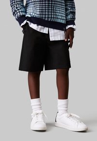 Pantaloni shorts neri con una vestibilità rilassata, abbinati a un maglione blu a fantasia, calze bianche alla caviglia e sneaker bianche con dettagli neri.