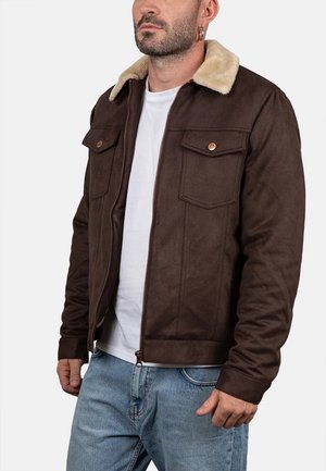 Uomo con giacca marrone con colletto in pelliccia beige sopra una maglietta bianca e jeans blu, in piedi davanti a uno sfondo neutro.
