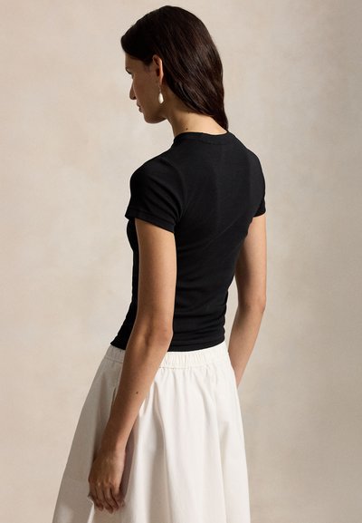 Γυναίκα με μακριά σκούρα μαλλιά, ντυμένη με εφαρμοστό μαύρο τοπ με κοντά μανίκια και λευκή φούστα με ψηλή μέση, πάνω σε απλό φόντο.