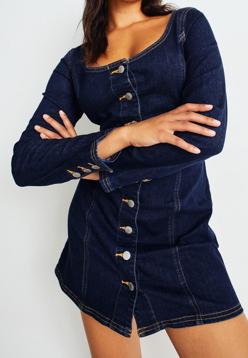 Femme portant une robe en denim bleu foncé avec boutons, manches longues, bras croisés, debout devant un fond clair uni.