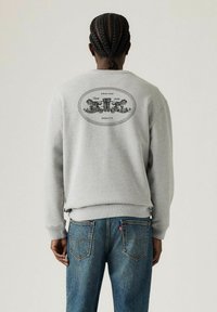 Sweat-shirt gris avec des poignets et un ourlet côtelés, présentant un design graphique noir au dos. Une paire de jeans en denim bleu avec des poches arrière visibles.
