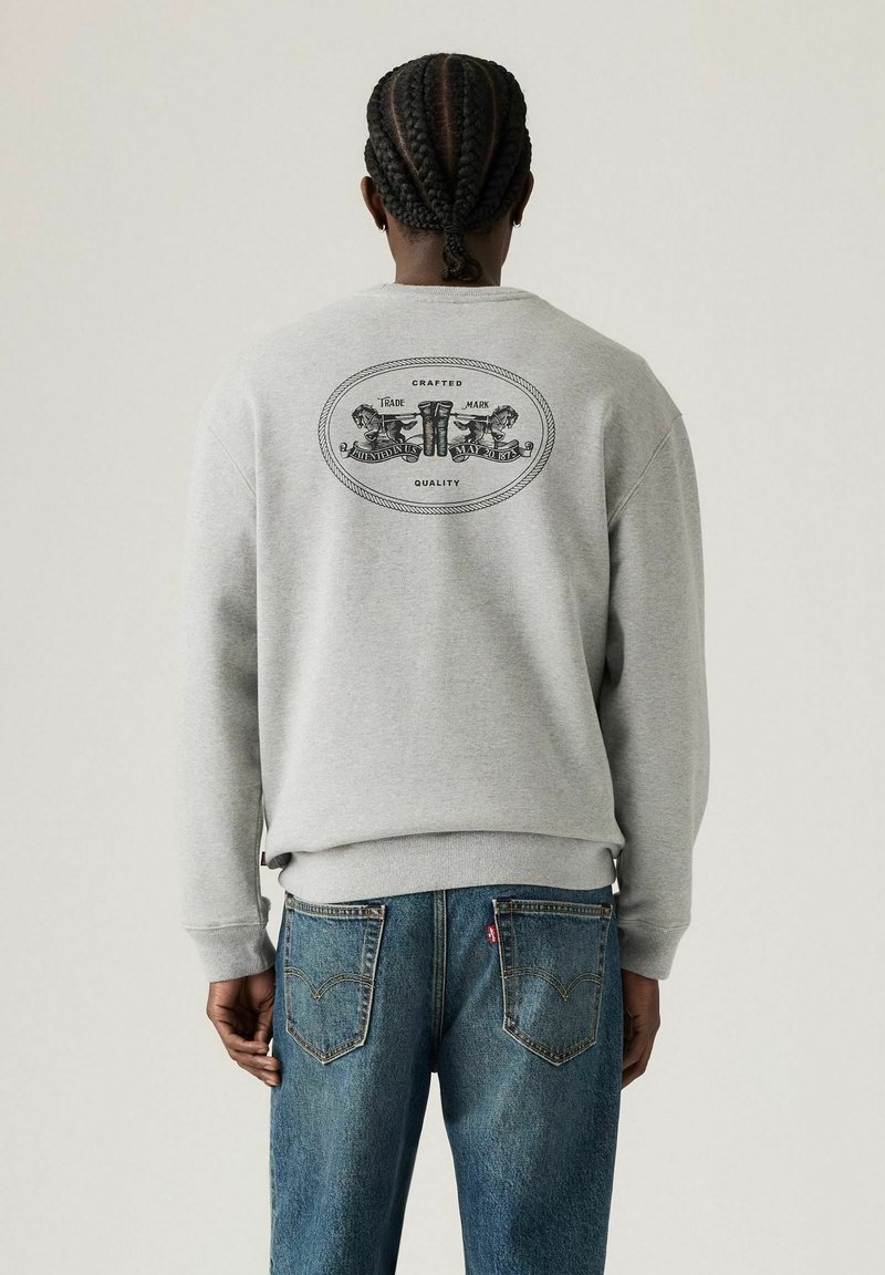 Sweat-shirt gris avec des poignets et un ourlet côtelés, présentant un design graphique noir au dos. Une paire de jeans en denim bleu avec des poches arrière visibles.