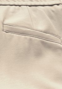 Lichtbeige stof met een diagonale ribbelstructuur en een stijlvolle, platte zakdetail. Het oppervlak is glad en netjes afgewerkt.