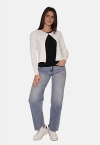 Cardigan lungo bianco con maniche lunghe sopra una maglietta nera, abbinato a jeans blu chiaro e scarpe da ginnastica bianche. Il cardigan presenta un collo rotondo e una silhouette aderente.