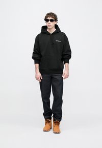 Sudadera negra con capucha, bolsillo delantero y cordón, vaqueros oscuros y botas color marrón claro. Estilo con gafas de sol oscuras. Diseño minimalista.