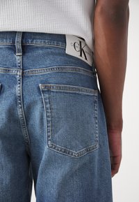 Blå denimjeans med en något urtvättad textur, med en vit Calvin Klein-etikett på midjebandet och två bakfickor.