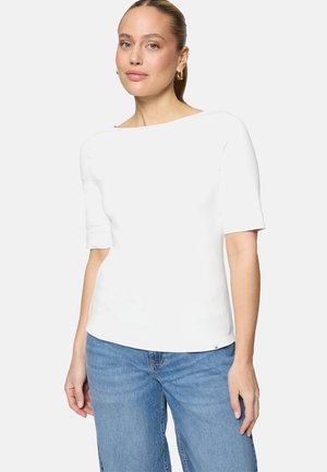 Frau mit hellem Teint und zurückgebundenem blondem Haar, die ein weißes Kurzarmshirt und blaue Jeans trägt, vor einem einfarbigen Hintergrund stehend.