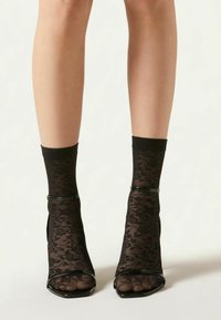 Černé sandály na podpatku s otevřenou špičkou, doplněné průsvitnými krajkovými kotníkovými ponožkami. Ponožky mají květinový vzor a sandály jsou ozdobeny černými pásky.