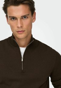 Pull marron à fermeture éclair avec col haut, tissu texturé et détail de fermeture éclair argenté, superposé à une chemise blanche.