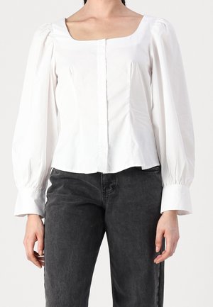 Femme portant un chemisier blanc à col carré avec de longues manches bouffantes et un jean noir, debout devant un fond clair.