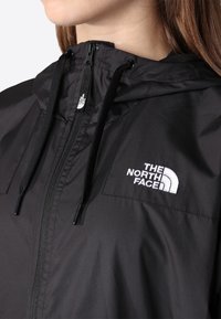 Gros plan d'une personne portant une veste noire North Face avec capuche, fermeture éclair visible, cordons et logo de la marque sur la poitrine.
