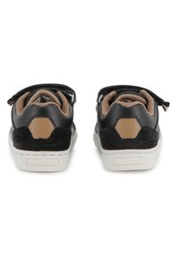 Baskets synthétiques noires avec une texture lisse, des accents beige et une semelle en caoutchouc blanche. Équipées d'une lanière Velcro pour la fermeture.