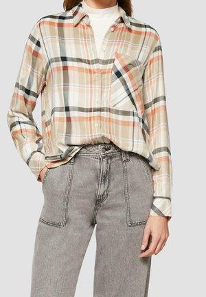 Kvinde iført beige, orange og sort ternet skjorte med knapper, der er stoppet ned i grå, højtaljede jeans med forlommer med lapper.