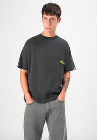 Black cotton t-shirt with a relaxed fit, featuring a small neon yellow logo on the left chest and short sleeves. Worn with gray jeans.
