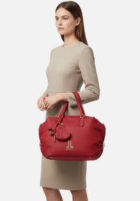 Borsa a tracolla in pelle rossa con due manici robusti, finiture in metallo color oro, una texture superficiale e un ciondolo a forma di cuore attaccato.