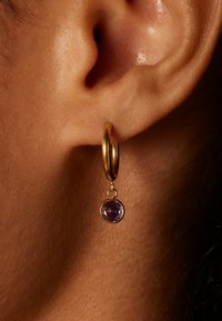 Gouden oorring met een hangende paarse edelsteen. De steen heeft een gefacetteerd oppervlak, wat een glanzend effect creëert. Close-up van een oor.
