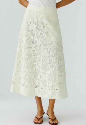 Frau trägt einen cremefarbenen Midirock aus floraler Spitze mit einem taillierten Bund, kombiniert mit braunen Flip-Flop-Sandalen vor einem schlichten Hintergrund.